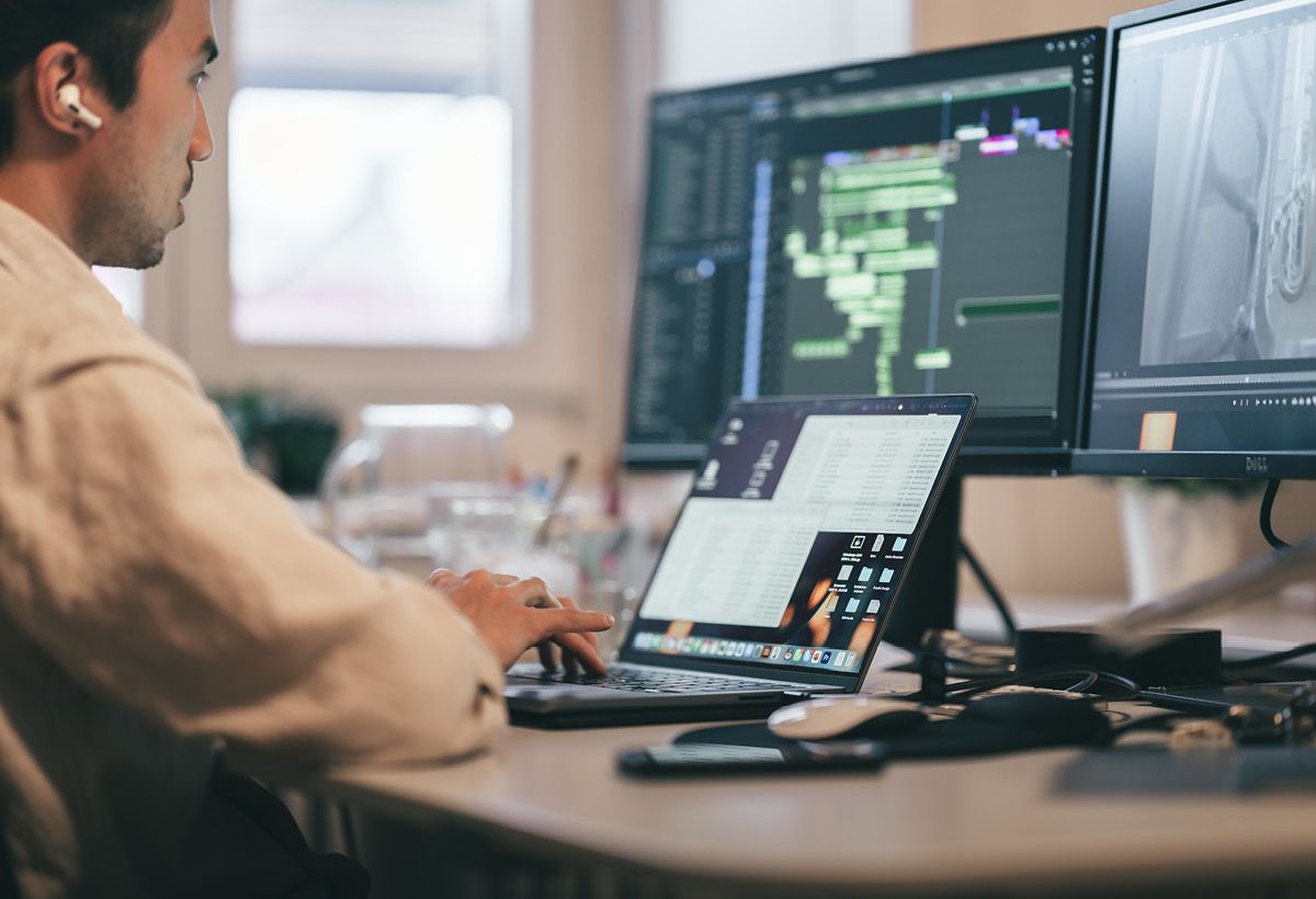 Kollege mit Ohrhörer arbeitet am Laptop und an zwei Bildschirmen